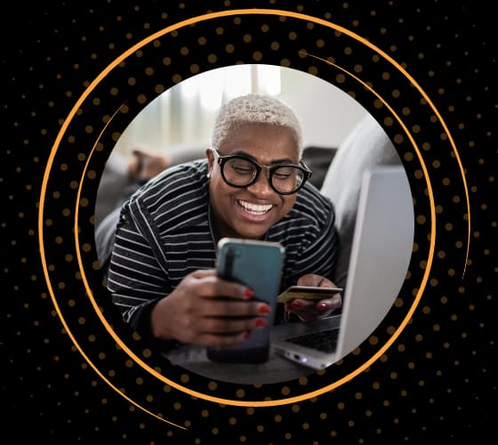 A person smiling while looking at a smartphone with a laptop open in front of them.